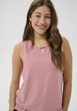 Jeune femme aux longs cheveux ondulés portant des boucles d'oreilles en forme d'anneau doré, un collier délicat et un haut rose ajouré sans manches, souriant à la caméra.