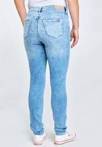 Isik, kes kannab helesiniseid kitsaid teksapükse tagataskutega ja valgeid tenniseid, seisab ühevärvilise tausta ees.