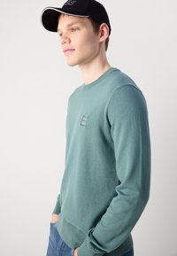 Young man wearing a teal crewneck sweater with a small logo and a black baseball cap, hands in pockets, looking sideways against a light background.