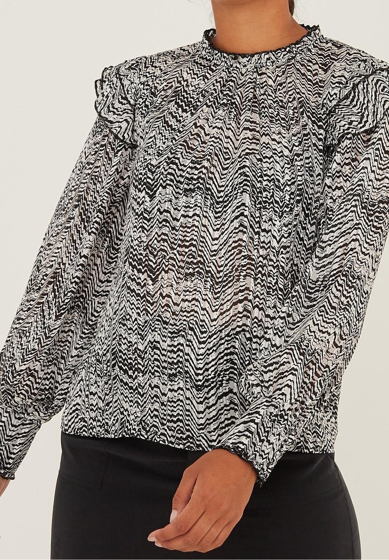 Vrouw met een zwart-wit zigzag patroon lange mouwen blouse met gerimpelde schouderdetails en een gegatherde halslijn.
