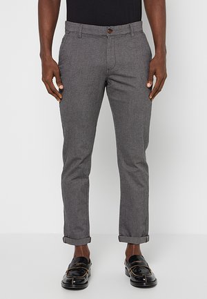 Homme portant un pantalon de costume gris slim avec des revers, des mocassins noirs à boucles dorées et une chemise noire rentrée, debout devant un fond uni.