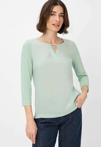 Zachtgroene top met een sleutelgat-halslijn, driekwartmouwen en een zachte textuur, gecombineerd met donkere denim broeken.
