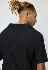 Chemise noire à manches courtes avec un motif en relief en forme de diamant, dotée d'un col et d'une coupe ample. La vue de dos montre une longueur d'épaule.