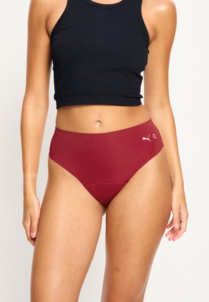 Femme portant un crop top noir sans manches et un sous-vêtement Puma bordeaux taille haute, debout devant un fond clair uni.