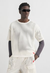 Homme portant des lunettes de soleil noires, une chemise en mesh blanche à manches courtes et un short en mesh assorti par-dessus une chemise à manches longues gris foncé, les mains dans les poches.