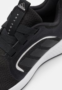 Zapatilla deportiva negra con parte superior de malla, tejido texturizado, cordones planos y tres distintivas franjas negras en el lateral. Suela blanca.