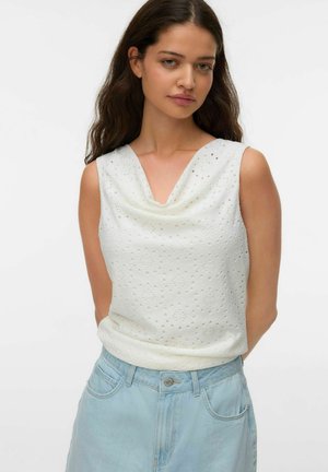 Ung kvinde med langt hår iført hvid ærmeløs øjlet-top og lyseblå højtaljede jeans mod en ensfarvet baggrund.