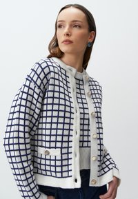 Gebreide cardigan met een marineblauw ruitpatroon op een witte achtergrond, met een ronde hals, knopen en zijzakken met decoratieve hardware.
