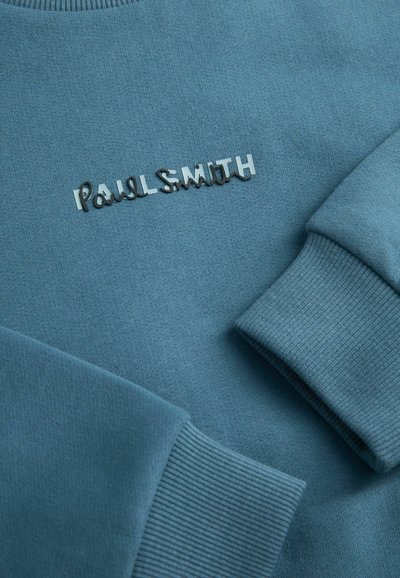 Sinine pusa, millel on soonikkoesed ja silmapaistev hõbedane "Paul Smith" logo, mis on õmmeldud ette, valmistatud siledast kangast.