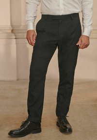 Schwarze, maßgeschneiderte Hose mit flachem Vorderteil, Gürtelschlaufen und schmaler Passform. Kombiniert mit polierten schwarzen eleganten Schuhen auf einem neutralen Hintergrund.