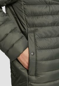 Olivgrüne Steppjacke mit gesteppten Abschnitten, ausgestattet mit einer Seitentasche mit einem schwarzen Druckknopf und einer glatten, glänzenden Stofftextur.