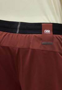 Κοντινή λήψη κόκκινων αθλητικών σορτς με μαύρη μέση που φέρει το λογότυπο της Ciele Athletics σε προσαρτημένη ετικέτα.