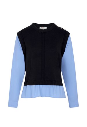 Zwarte gebreide mouwloze trui met drie decoratieve knopen op de schouder, gedragen over een lichtblauw bloesje met lange mouwen en omslagboorden.