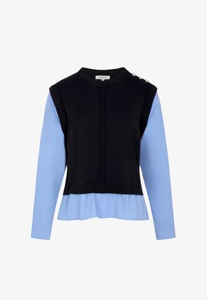 Zwarte gebreide mouwloze trui met drie decoratieve knopen op de schouder, gedragen over een lichtblauw bloesje met lange mouwen en omslagboorden.