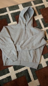 Ανοιχτό γκρι φούτερ με φερμουάρ, μπροστινές τσέπες και κουκούλα, απλωμένο επίπεδα σε μοτίβο καφέ, πράσινου και κρεμ χαλί.