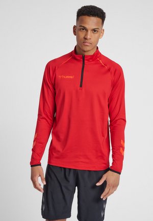 Rotes langärmliges Sportshirt mit schwarzem Reißverschluss und orangefarbenen Akzenten. Das Material wirkt glatt. Kombiniert mit schwarzen Shorts. Minimales Logo auf der Brust.