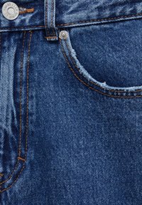Gros plan d'une poche de jean en denim bleu avec une surpiqûre orange, des rivets métalliques et une boucle de ceinture en tissu visible en haut.
