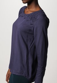 Mörkblå långärmad blus med texturerad tyg och en blommig spetsdetalj runt halsen. Har en avslappnad passform och gathered ärmar.