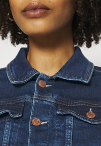 Dark blue denim jacket with a collared neck, copper buttons, and distressed stitching details. Close-up view focusing on upper section.