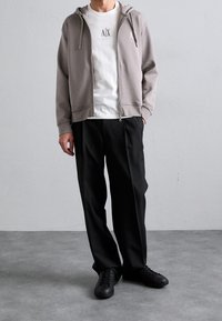 Felpa con cappuccio grigia con zip e logo sul petto, indossata sopra una T-shirt bianca. Abbinata a pantaloni neri e sneakers nere, in piedi su una superficie grigia.