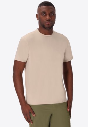 Uomo con capelli corti e barba che indossa una maglietta beige a maniche corte e pantaloni verde oliva, in piedi davanti a uno sfondo chiaro e semplice.