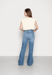 Camisa oversized bege com mangas em forma de capa, combinada com jeans claros com pernas largas, apresentando bolsos traseiros em forma de coração e uma textura de denim suave.