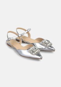 Zapatos planos metálicos plateados con puntera puntiaguda, correas ajustables en el tobillo y decoración de cristal en la parte delantera. Textura suave, tacón bajo.