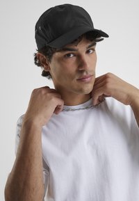 Casquette noire avec une couronne structurée et un bord incurvé, portée par une personne en chemise blanche. La casquette possède des œillets de ventilation et un design épuré.