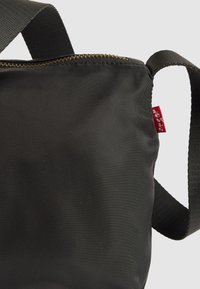 Zwarte nylon schoudertas met verstelbare band, messing rits en klein rood stoffen label aan de zijkant op een effen achtergrond.