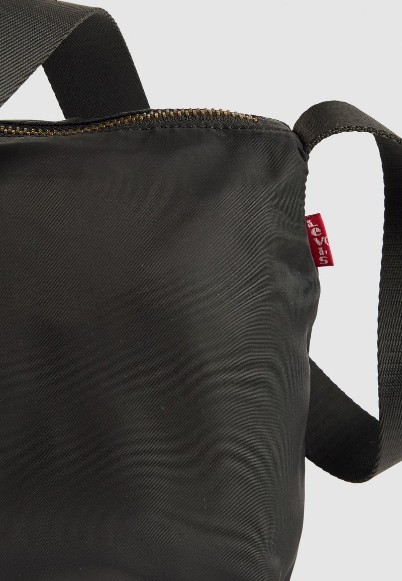 Bolso cruzado de nylon negro con correa ajustable, cremallera de latón y pequeña etiqueta de tela roja en el lateral, sobre un fondo sencillo.