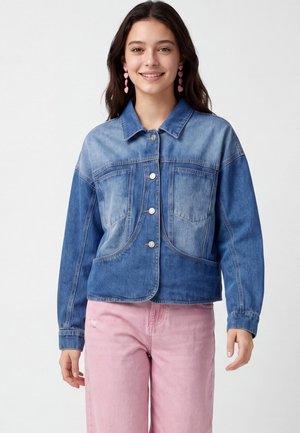 Jonge vrouw die glimlacht en een tweekleurige blauwe spijkerjas, roze hartvormige oorbellen en lichtroze broek draagt, staande tegen een effen achtergrond.