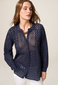 Dunkelbluse mit Knopfleiste, die aufwändige Laserschnittmuster, einen runden Ausschnitt und lange Ärmel mit knöpfbaren Manschetten aufweist.