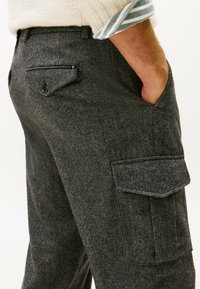 Hombre vestido con pantalones cargo de espiga gris oscuro, con bolsillos de solapa abotonados, mano en el bolsillo lateral, y un suéter crema con puños de manga a rayas.