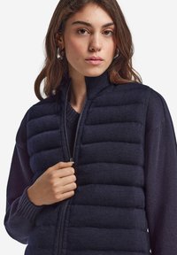 Mörkblåt isoleringsväst med quiltad textur; hög krage och dragkedja; bärs över en mörk tröja; hand som greppar dragkedjan.