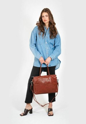 Brown leather handbag with two handles, adjustable shoulder strap, and front pocket, paired with a denim shirt and black pants.