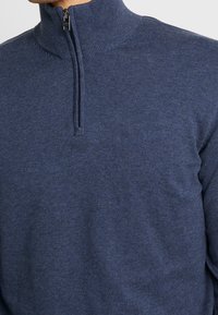 Homme portant un pull à demi-fermeture éclair bleu avec un col côtelé et une texture subtile, montrant des détails de la poitrine et du haut du bras.