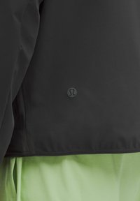 Närbild av fållen på en svart jacka med en liten rund logotyp ovanför ljusgrön tyg med subtilt rutmönster.