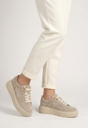 Füße in beige Wildleder-Plateau-Sneakers mit cremefarbenen Schnürsenkeln, kombiniert mit hochgekrempelten, elfenbeinfarbenen Hosen auf hellem Hintergrund.