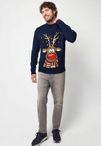 Donkerblauwe trui met een feestelijk rendierontwerp; heeft kleurrijke verlichting, een rode neus en een gestreepte sjaal. Ggedragen met grijze jeans en donkere schoenen.