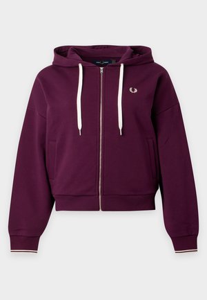 Femme portant un sweat à capuche zippé bordeaux, une chemise blanche, un pantalon large beige et des chaussures noires, debout les mains dans les poches sur un fond blanc uni.