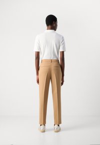 Tan tailored pants with a slim fit, featuring back pockets and a smooth texture. Paired with a plain white polo shirt and white sneakers.