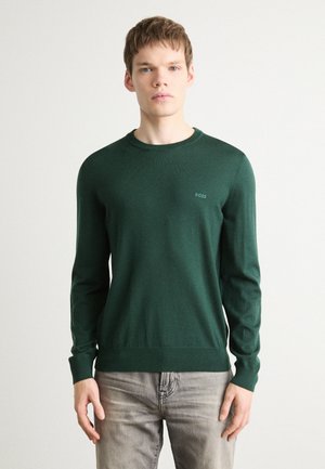 Maglione verde a maglia con scollo rotondo, polsini e orlo a costine, caratterizzato da un piccolo logo sul lato sinistro del petto. Indossato sopra jeans grigi chiari.