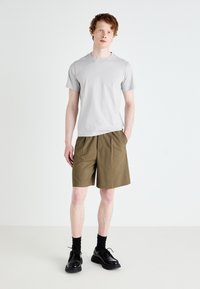 Grijze katoenen crew neck t-shirt gecombineerd met kaki geplooide shorts. Het model draagt zwarte veterschoenen en zwarte sokken. Eenvoudig, minimalistisch ontwerp.
