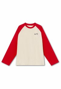 Koszulka raglan z długim rękawem, czerwonymi rękawami i kołnierzykiem, beżowym korpusem oraz małym czarnym logo w kształcie spirali po lewej stronie klatki piersiowej.