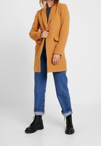 Manteau de couleur moutarde en tissu doux, avec un col à revers cranté, deux poches avant et une coupe ajustée. Porté avec un jean bleu et des bottes noires.