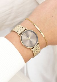 Montre en métal doré avec un bracelet texturé et un cadran gris. Le cadran présente des index horaires dorés et un texte de marque discret sous la position de 12 heures.