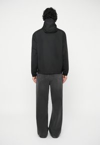 Asmuo su garbanotais plaukais, vilkintis juodą striukę su kapuce ir laisvas tamsiai pilkas džinsus, stovi atsukęs nugarą prieš paprastą baltą foną.