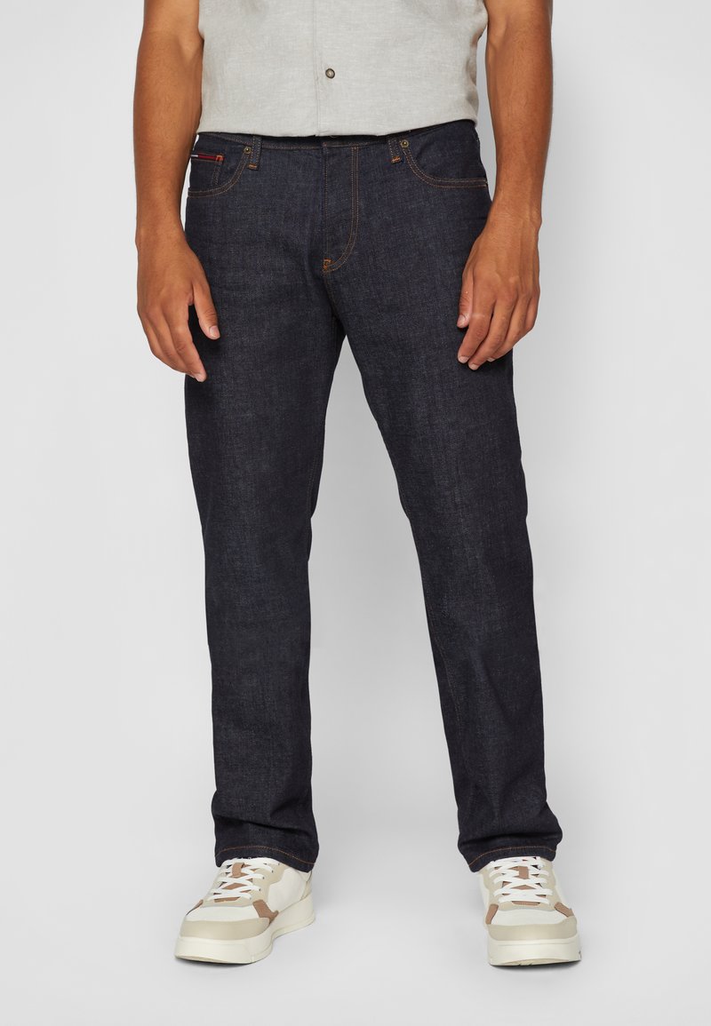 Man in een donkerblauwe rechte jeans en witte sneakers, staande tegen een effen grijze achtergrond.