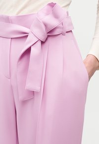 Παντελόνι σε λιλά με υψηλή μέση, εμπρός πιέτες και πλατύ υφασμάτινο ζωνάκι δεμένο σε φιόγκο, συνδυασμένο με κρεμ μακρυμάνικη μπλούζα και ένα χέρι στην τσέπη.