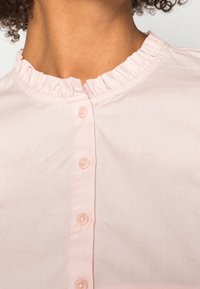Camisa de botones rosa con cuello rizado, que cuenta con cinco botones redondos y una textura de tejido suave y ligero.
